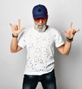 Old brutal man in designer t-shirt, blue jeans and baseball cap shows rock with both hands stylish fashionable men