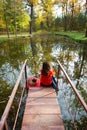 An old camera lies on a suitcase on a red cage. A girl sits on a wooden bridge by the lake