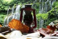 Oil lantern with autumn leaves on blurry waterfall background in the forest.