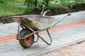 An old dirty metal wheelbarrow with an orange wheel on the sidewalk. Used construction tool, side view. Agricultural Royalty Free Stock Photo