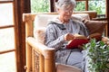 Old elderly woman sitting at home on armchair reading a book wearing a warm sweater and eyeglasses. Comfortable living room, Royalty Free Stock Photo