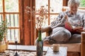 Old elderly woman sitting at home on armchair reading a book wearing a warm sweater and eyeglasses. Comfortable living room, Royalty Free Stock Photo