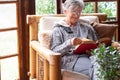 Old elderly woman sitting at home on armchair reading a book wearing a warm sweater and eyeglasses. Comfortable living room, Royalty Free Stock Photo