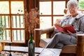 Old elderly woman sitting at home on armchair reading a book wearing a warm sweater and eyeglasses. Comfortable living room, Royalty Free Stock Photo