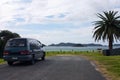 An old family car /backpacker van parked by the sea on a tarmac road, a palm tree on the right, in Northland in New Zealand