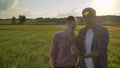 Old farmer discussing new project with his successor and holding tablet, standing on wheat field during beautiful sunset