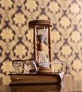 Old fashioned attributes concept. Hourglass, old book and eyeglasses on wooden table, pattern background. Sand falling