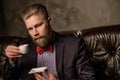 Old-fashioned bearded man sitting in comfortable leather sofa with cup of coffee on gray. Royalty Free Stock Photo