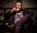 Old-fashioned bearded man sitting in comfortable leather sofa with cup of coffee isolated on gray. Royalty Free Stock Photo