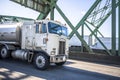 Old fashioned cab over big rig semi truck transporting fuel in tank semi trailer running on the truss arched Interstate Bridge