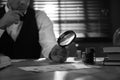 Old fashioned detective with magnifying glass working at table in office, closeup. Black and white effect Royalty Free Stock Photo