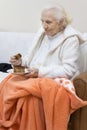 Old gray-haired woman in a white dressing gown sits on a sofa and eats a cake from a saucer.