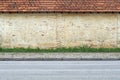 Old grunge yellow brick wall with terracotta tiled roof. A stripe of grass, concrete tiled sidewalk and asphalt road in front.