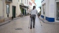 Old man with two walking sticks or crutches walking with little steps on street.