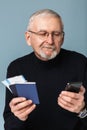 Old handsome smiling man with gray hair and beard in eyeglasses and sweater holding tickets and passports in hand while Royalty Free Stock Photo