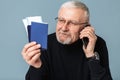 Old handsome smiling man with gray hair and beard in eyeglasses and sweater holding tickets and passports in hand while Royalty Free Stock Photo