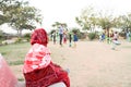 An old indian lady sitting on the bench and watching children playing in an open gym in a park