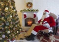 Old kind bearded Santa Claus sitting at armchair in his cozy home room near Christmas tree, fireplace, xmas stocking and big gift Royalty Free Stock Photo