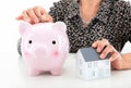 An old lady holds a small house model in one hand and puts a coin into the piggy bank in the other hand