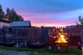 An old lonely man sits sadly on a bench at a village house behind a fence looking into the fire of a barbecue fire