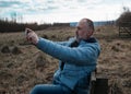 Old man in a blue coat with the mobile phone sitting on the bench in a field