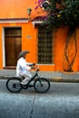 Old man in cowboy hat cycling along quiet street past colorful orange house