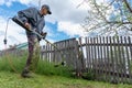 An old man with an electric lawn mower trimmer cuts fresh green grass by the fence wearing glasses and a cap Royalty Free Stock Photo