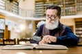 Old man with gray beard, in glasses, vintage clothes, reading a book in the ancient library, using magnifying glass