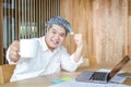 The old man holding a white coffee cup sitting on a brown wooden table with a notebook computer Royalty Free Stock Photo