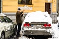 Old man, male in winter coat cleaning, shoveling driveway, street from covered falling snow with shovel, residential houses, cars