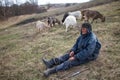 An old man in messy clothes is sitting on a hill and herding a flock of his own goats against the backdrop of withered nature Royalty Free Stock Photo