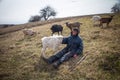 An old man in messy clothes is sitting on a hill and herding a flock of his own goats against the backdrop of withered nature Royalty Free Stock Photo