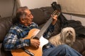 Old man playing acoustic guitar and sitting on sofa with little white dog. Royalty Free Stock Photo