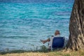 The old man sits on a folding chair next to a tree and reads the book with sea water in background Royalty Free Stock Photo