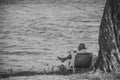 The old man sits on a folding chair next to a tree and reads the book with sea water in background. Black and white Royalty Free Stock Photo