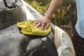 An old man uses a yellow washcloth to clean the hood of his black hatchback. A car owner cleaning and taking care of his auto at
