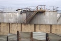 Old metal industrial storage tanks with rusty inspection ladders and valves surrounded by a corrugated steel fence and wall