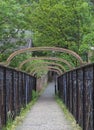 Old metal pedestrian footbridge crossing a railway line leading to a narrow alley surrounded by trees and rustic buildings