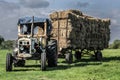 Old neglected blue tractor pulling a triler of hay in a field at harvest time