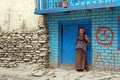 Old Nepalese woman in a traditional dress stands near the blue wall, on the street in the village of Kagbeni of the Kingdom Mustan