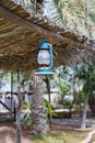 An old oil lamp hangs on a canopy of palm leaves, against the backdrop of a tropical village and palm trees
