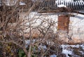 An old ramshackle house with a boarded-up window, a leaning chimney, and a felled plum tree. Sunny day, snow on the ground