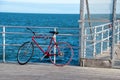 An old red bicycle leaning against and chained to a fence and hand rail on a wooden pier with the blue ocean in the background Royalty Free Stock Photo