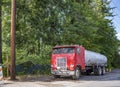 Old red big rig cabover semi truck with tank trailer standing on the road shoulder on the green trees background