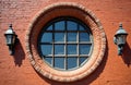 Old red brick wall features large round window with intricate brickwork frame. Window multiple glass panes reflecting light. Two Royalty Free Stock Photo