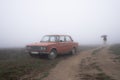 Old red retro car stands in fog near the dirt road, girl in white coat walks under umbrella on the background Royalty Free Stock Photo