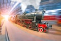 An old retro locomotive train railroad with steam and smoke clubs arrives at the passenger covered station under the roof. Royalty Free Stock Photo