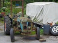 Old rusting abandoned tractor next to a dirty tarpaulin covered trailer and junk in a farmyard