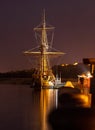 An old sailing ship, at night, in the light of lanterns. The night city, the embankment, reflections in the black water.