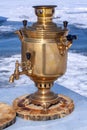 An old samovar stands on a cut of a tree trunk on an ice plate on a background of snow. Royalty Free Stock Photo